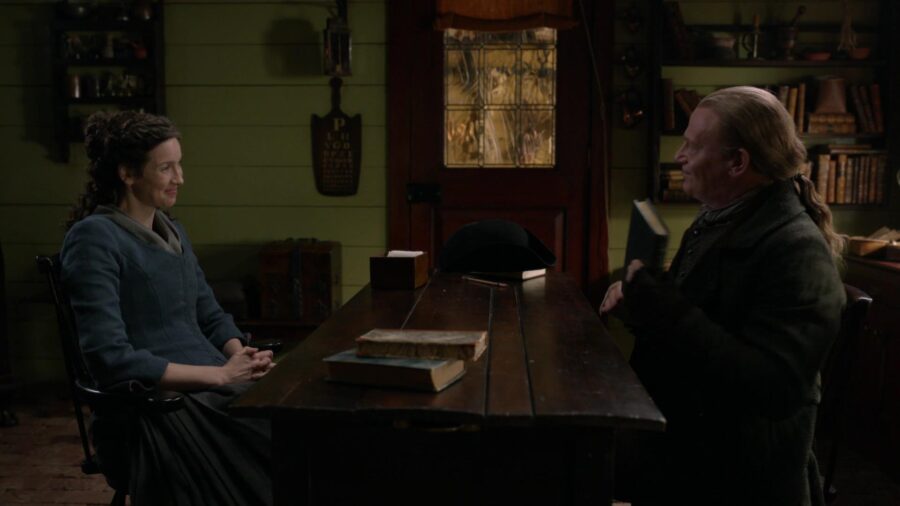 Claire and Tom sit at a table with books. Tom is holding one in his hand. 